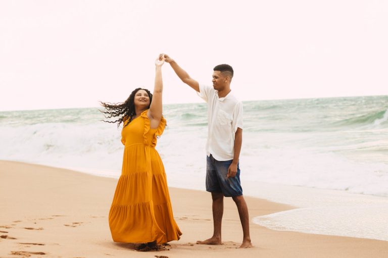 Dos personas disfrutando de las clases de baile latino en la playa, con la mujer en un vestido amarillo fluido y el hombre con camisa blanca y shorts azules, representando la esencia del baile latino en la arena