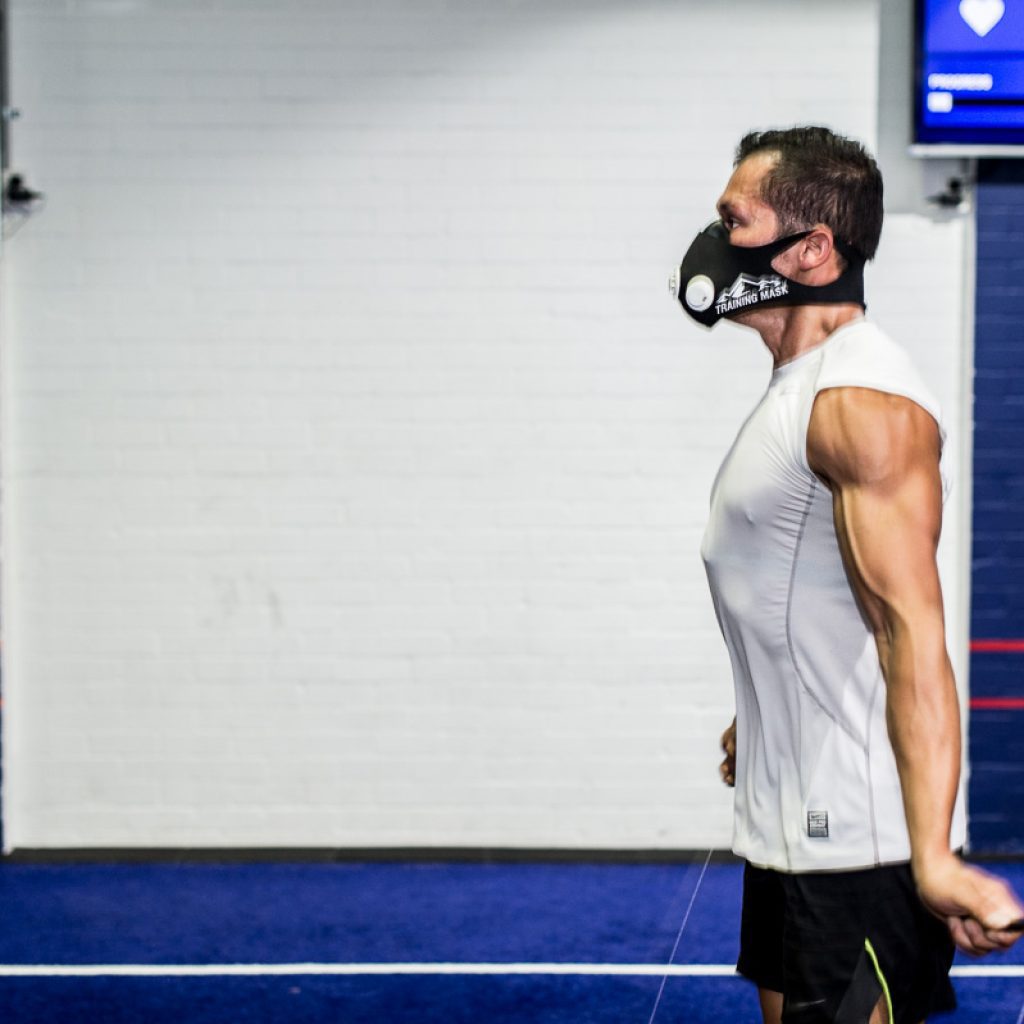Hombre realizando ejercicios cardio en un gimnasio con una máscara de entrenamiento de resistencia.